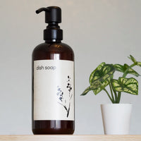 amber glass bottle with black satin steel pump on a gray background with a Japandi Dish Soap label and a small plant.