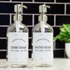 Clear glass apothecary soap dispensers with labels for hand soap and dish soap with stainless steel pumps and waterproof labels in a black tile kitchen.