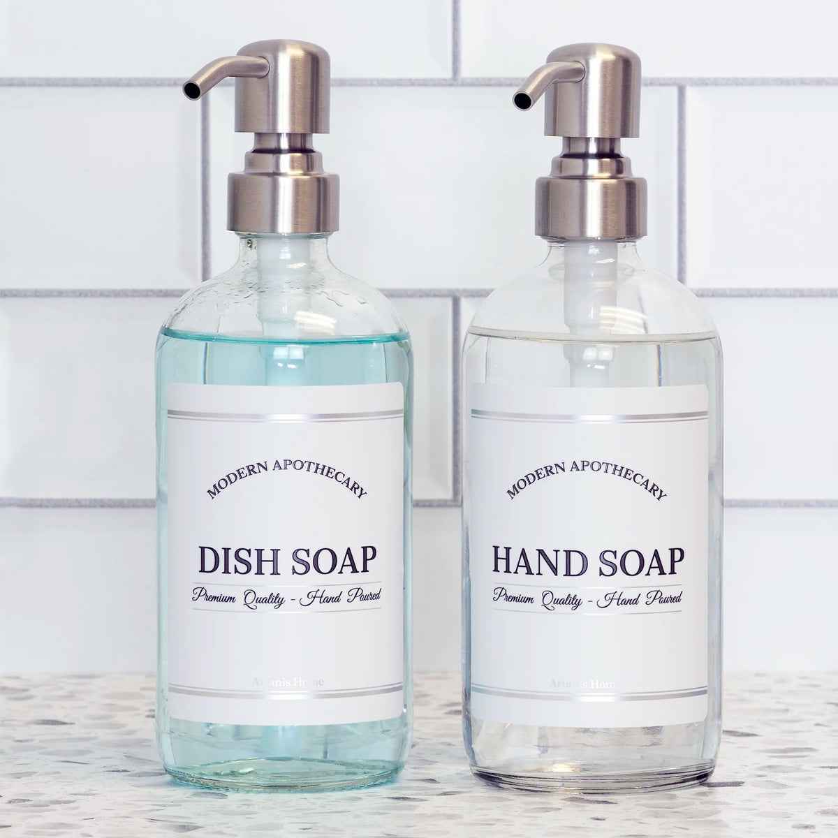 Clear glass hand soap dish soap set of soap dispensers with pretty labels and stainless steel pumps on a white tile kitchen renovation countertop. Labels are coordinated to declutter.