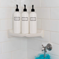 Tall white plastic set of shower bottles with black pumps and silkscreen print for shampoo, conditioner, and body wash, on shower shelf.