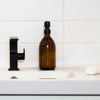 16 oz amber glass apothecary bottle with black satin steel pump, sitting at a bathroom sink.